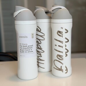 May include: Three white Owala water bottles with gray lids. One bottle has a label with text, "Hi there. I'm leak proof. I'm insulated. And I have a built-in straw." The other two bottles have names in silver script: "Anapelle" and "Dalila."