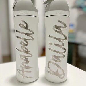 May include: Two white Owala water bottles with gray lids. One bottle has the name "Anabelle" in silver script, and the other has "Dalila". The bottles are on a white surface.