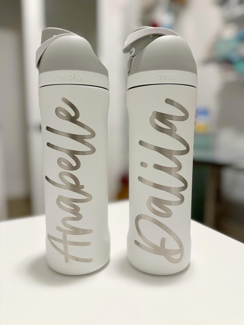 May include: Two white Owala water bottles with gray lids. One bottle has the name "Anabelle" in silver script, and the other has "Dalila". The bottles are on a white surface.