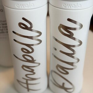 May include: Two white Owala water bottles with gray lids. The bottles have silver cursive lettering that reads "Anabelle" and "Dalila". The bottles are on a white surface.