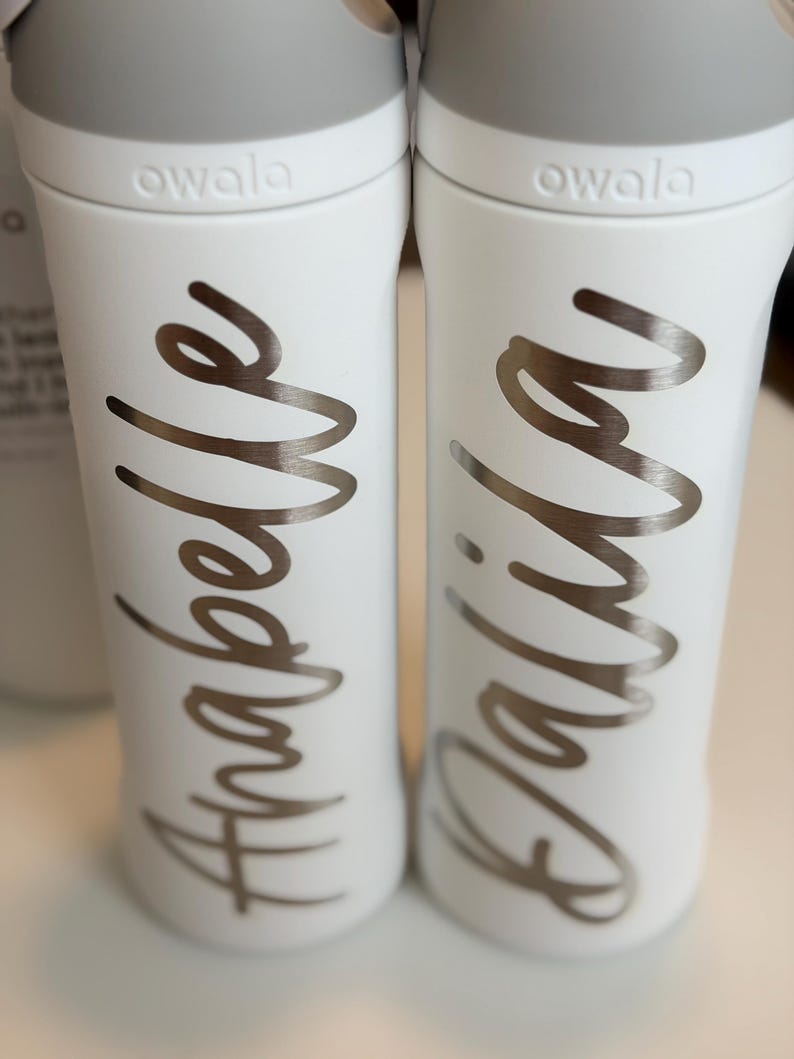 May include: Two white Owala water bottles with gray lids. The bottles have silver cursive lettering that reads "Anabelle" and "Dalila". The bottles are on a white surface.