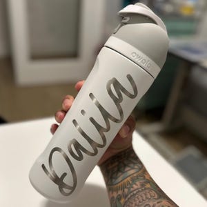 May include: A white water bottle with a gray lid and the name "Daila" in a silver script. The bottle has the brand name "owala" printed on it. The bottle is held in a hand.