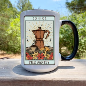 May include: A large white coffee mug with a black handle. The mug features a tarot card design with a brown coffee maker, a cup of coffee, and the text "THE SANITY". The design includes flowers and celestial elements.