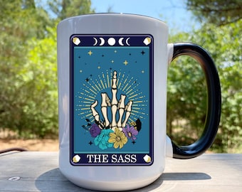 May include: White ceramic mug with a black handle and rim. The mug features a tarot card design with a skeleton hand giving the middle finger, surrounded by flowers and the text "THE SASS". The background is a teal color with stars and moon phases.