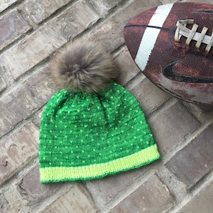 May include: A green and yellow knitted hat with a brown faux fur pom pom. The hat is sitting on a brick surface next to a brown football.