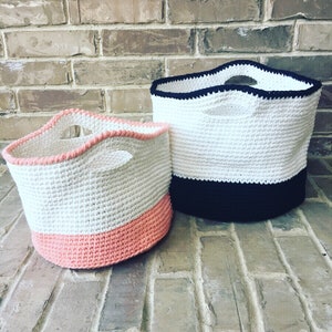 May include: Two crocheted storage baskets with handles. The larger basket is white with a black bottom band. The smaller basket is white with a pink bottom band.