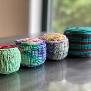 Peut inclure: Quatre petits contenants ronds tricotés, alignés. Les contenants sont de couleurs variées, dont le vert, le bleu et le gris, avec des motifs rayés et à motifs. Les contenants semblent faits à la main et sont posés sur une surface réfléchissante.