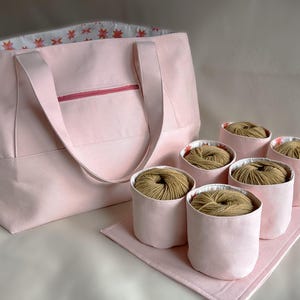 Könnte beinhalten: Eine rosa Canvas-Tragetasche mit Reißverschlussfach und sechs rosa Stoffbehältern gefüllt mit Garn. Die Behälter stehen auf einer rosa Stoffmatte.