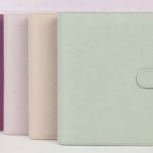 May include: A set of four rectangular planners with textured covers in shades of purple, light gray, beige, and mint green. Each planner features a vertical spine and a matching tab closure on the right side, arranged in a row from left to right.