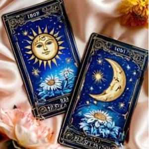 May include: Two tarot cards with celestial designs. One features a smiling sun, the other a crescent moon. Both cards have a blue background with white flowers and gold accents. The cards are on a satin surface.