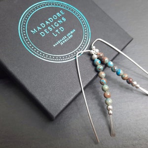 May include: A pair of silver earrings with a long, thin wire design. Each earring features a strand of small, round, multi-coloured beads. The beads are a mix of brown, blue, and green.
