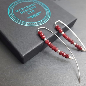 Puede incluir: Un par de pendientes de plata con cuentas de piedras preciosas rojas. Los pendientes son largos y colgantes, con un diseño de alambre curvo.