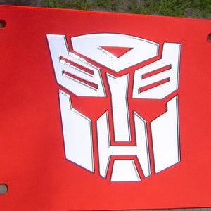 May include: A red metal license plate with a white 3D Transformers logo. The logo is a stylized robot face with a triangular head and a rectangular body.