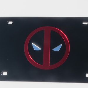 May include: Black metal license plate with a red Deadpool logo. The logo is a red circle with a white outline and a black Deadpool mask inside. The mask has two white eyes.