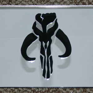 Puede incluir: Una placa de matr&iacute;cula de metal blanco con un s&iacute;mbolo mandaloriano negro. El s&iacute;mbolo es una calavera con cuernos y una l&iacute;nea vertical en el centro.