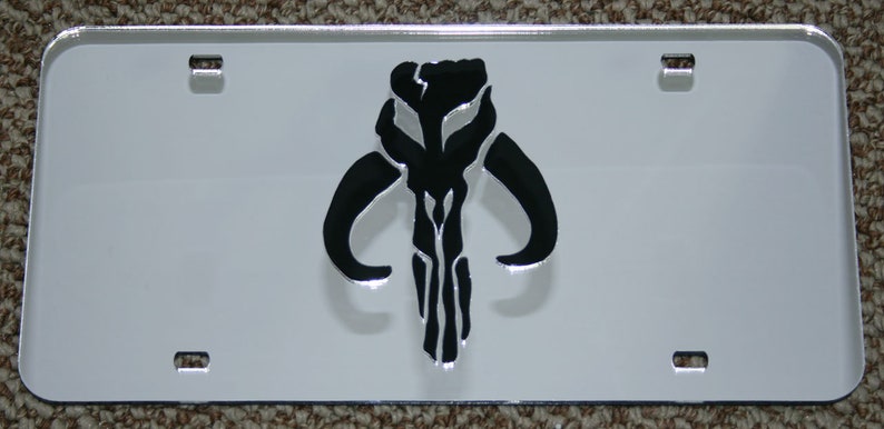 Puede incluir: Una placa de matr&iacute;cula de metal blanco con un s&iacute;mbolo mandaloriano negro. El s&iacute;mbolo es una calavera con cuernos y una l&iacute;nea vertical en el centro.