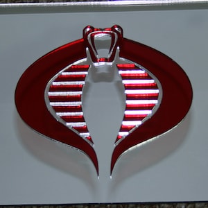 May include: A silver license plate featuring a red cobra emblem. The emblem is a stylized cobra head with a red outline and white stripes. The plate has four mounting holes.