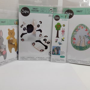 May include: A set of five die-cut craft supplies featuring a panda bear, a bunny rabbit, and a tree with birds. The die-cuts are packaged in clear plastic bags with the Sizzix brand logo and the number of dies included. The packaging also includes the text "Thinlits Fold-a-Long" and "Thinlits".