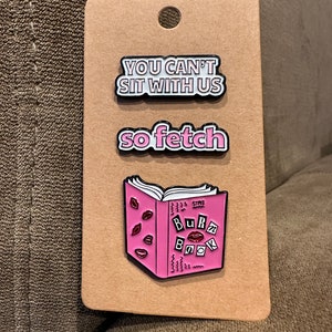 Puede incluir: Tres pines de esmalte sobre un fondo de cartulina marrón. Los pines son de color rosa y blanco con contornos negros. El pin superior dice "You can't sit with us". El pin del medio dice "so fetch". El pin inferior es un libro rosa con las palabras "Burn Book" en la portada.