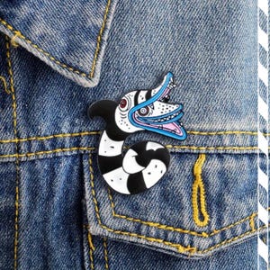 May include: A black and white enamel pin featuring a cartoon monster with a large mouth and teeth. The monster has a blue tongue and is wearing a black and white striped pattern.