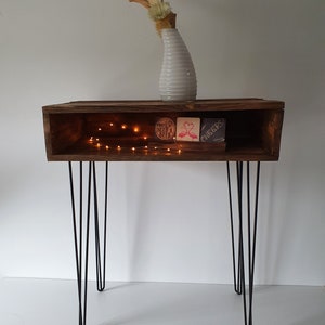 Könnte beinhalten: Ein Holz-Beistelltisch mit einem Regal und Haarnadelbeinen. Das Regal hat ein kleines rundes Schild mit der Aufschrift "You'll Never Be Alone" und eine Karte mit einem rosa Flamingo mit dem Text "Cheers". Auf dem Tisch steht eine weiße Vase mit getrockneten Blumen.