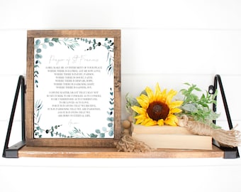 May include: A framed print with a white background and a blue and green watercolour design of eucalyptus leaves. The print features the text "Prayer of St. Francis" and the prayer itself. The frame is brown wood with a white mount.