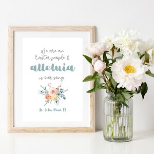 May include: A framed print with a white background and a quote in blue text that reads "We are an Easter people & alleluia is our song. -St. John Paul II-". The quote is surrounded by a watercolour floral illustration.