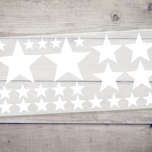 Könnte beinhalten: Eine Bogen mit weißen Stern-Aufklebern auf einem Holz-Hintergrund. Die Sterne sind in verschiedenen Größen und in einem zufälligen Muster angeordnet.