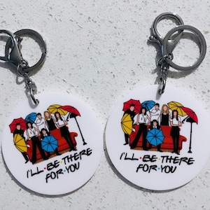 May include: Two round white keyrings with silver key rings. Each keychain features a colourful illustration of the Friends TV show characters sitting on a red couch with umbrellas behind them. The text "I'll Be There For You" is printed below the illustration.