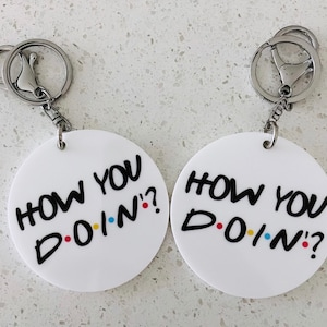 May include: Two white round keyrings with the phrase "How You Doin'?" in black text with colourful dots. Each keyring has a silver metal ring.
