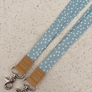 May include: Two pale blue lanyards with white polka dots. Each lanyard features a silver-coloured clasp and a small, rectangular, tan leather tag with the text "Lanyards by Zoe".