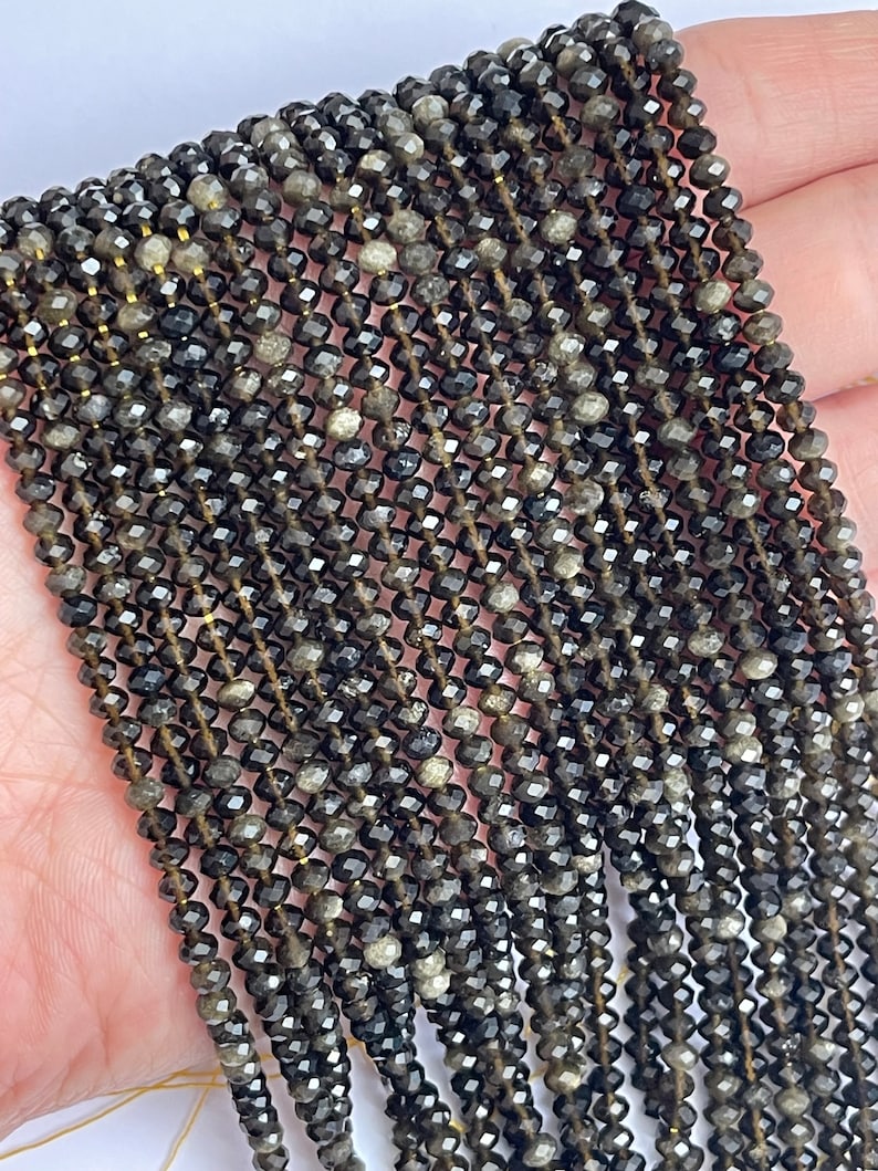 May include: A strand of black and gold gemstone beads. The beads are faceted and have a shiny finish. The beads are strung on a thin wire.