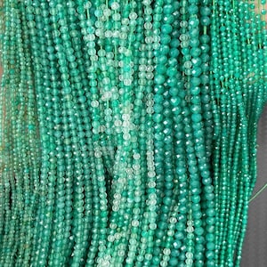 May include: Multiple strands of faceted round beads in various shades of green, ranging from light mint to deep emerald. The beads are strung on thin green thread, creating a dense, vertical arrangement of small, reflective, circular gemstone components.