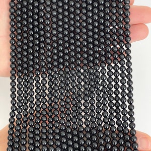 May include: Close-up of strands of shiny, black, spherical beads. The beads are consistent in size and strung together. The beads are held in a hand against a white backdrop. These beads are likely for jewellery making.