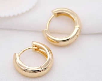 May include: Two gold-coloured hoop earrings. The earrings have a smooth, polished surface and a hinged closure. The hoops are a medium size and have a rounded shape. The earrings are displayed on a white surface.