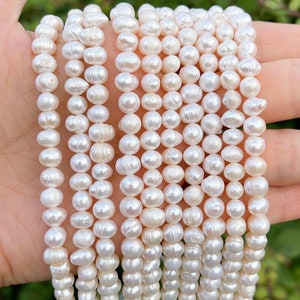 Könnte beinhalten: Eine Strähne weißer Süßwasserperlen mit einer glatten, leicht unregelmäßigen Oberfläche. Die Perlen sind auf einem durchsichtigen Faden aufgefädelt und haben einen Durchmesser von etwa 6-7 mm.