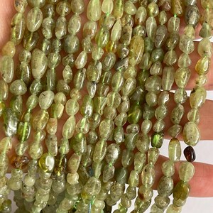 May include: Strands of olive green gemstone beads. The beads are oval and irregular in shape, strung together with green thread. The beads vary in colour from light to dark green, with some having a translucent quality. The beads are a natural stone.