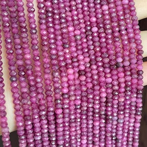May include: Multiple strands of small, round, faceted ruby beads in a deep magenta-pink color. Each bead features a reflective, multi-faceted surface that catches the light, arranged in long, vertical, parallel lines on a thin string.