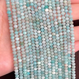 May include: A strand of faceted round beads in varying shades of light blue, teal, and pale green. The small, semi-translucent stones are strung together on a thin blue thread, displaying natural colour variations and a reflective, multi-faceted surface.
