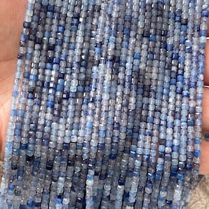 Könnte beinhalten: Eine Strähne blauer Kyanit-Perlen, jede Perle hat eine quadratische Form mit einer facettierten Oberfläche. Die Perlen sind eine Mischung aus hellblauen und dunkelblauen Farbtönen.