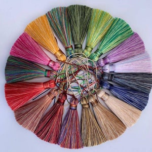 May include: A collection of 15 colourful silk tassels, each with a different colour combination. The tassels are arranged in a circle, with the tips of the tassels pointing outwards. The tassels are all about 7.5 cm long.