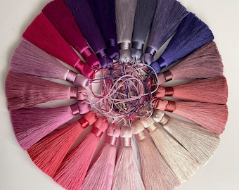 May include: A circular arrangement of tassels in various shades of pink, red, purple, and grey. Each tassel has a silky appearance and is tied with a matching ribbon. The image is taken from above, showcasing the array of colours and textures.