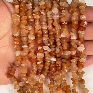 May include: A collection of small, irregular-shaped gemstones in shades of orange, peach, and cream. The polished beads are strung together, creating a textured display. These gemstones are likely used for jewellery making.