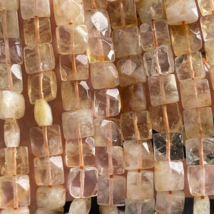 May include: Close-up of strands of citrine gemstone beads. The beads are square-cut and faceted, with a translucent, golden-yellow hue. Each bead is strung on a thin, orange-coloured cord, creating a textured, natural look. The beads are arranged closely together.