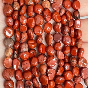 May include: A strand of red jasper gemstone beads, tumbled and polished into smooth, pebble-like shapes. The beads are a rich, reddish-brown colour with subtle variations in tone and texture.