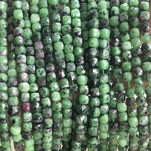 Puede incluir: Una hebra de cuentas de piedra preciosa facetadas de color verde y negro. Las cuentas son de forma cuadrada y tienen una textura rugosa y natural.