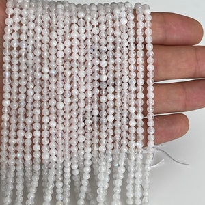 May include: Strands of small, round, white beads. The beads are strung together, creating a delicate and shimmering effect. The beads are held in a hand against a white background. The beads appear to be made of a translucent material.