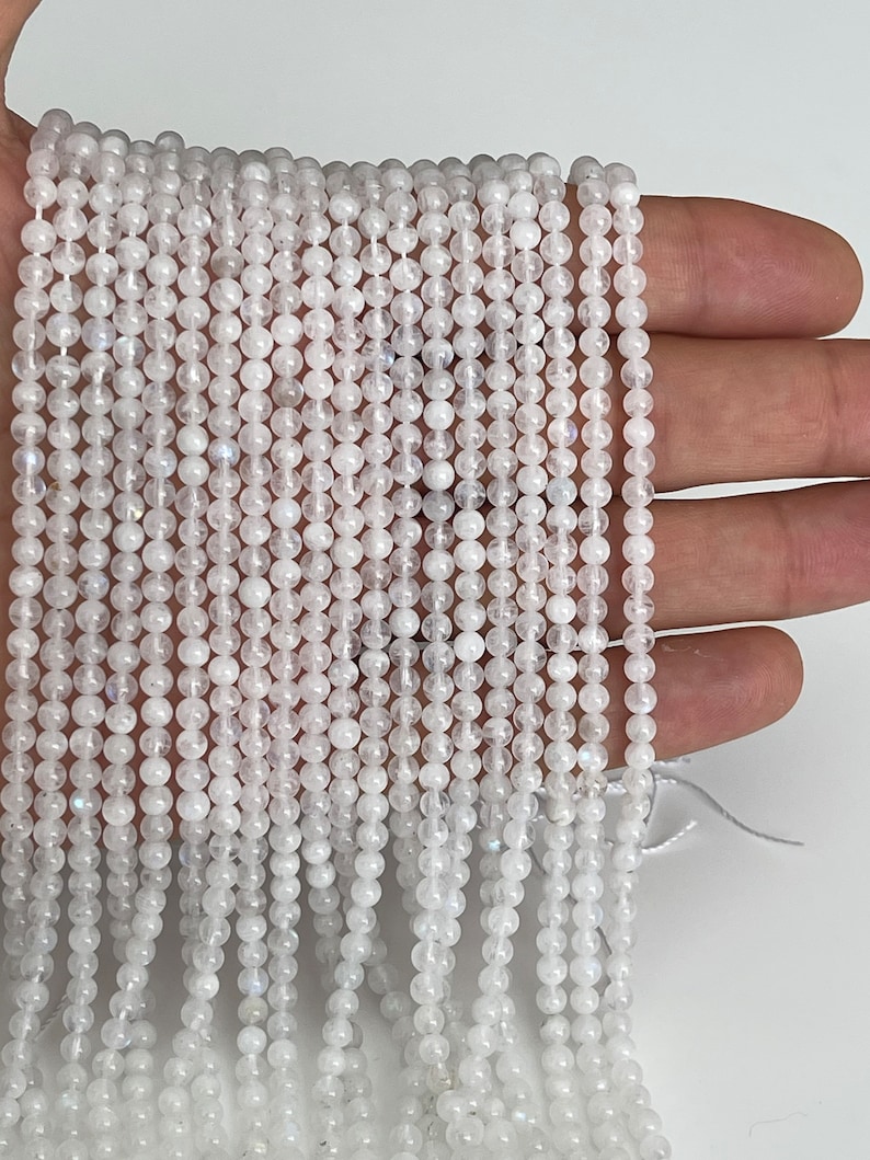 May include: Strands of small, round, white beads. The beads are strung together, creating a delicate and shimmering effect. The beads are held in a hand against a white background. The beads appear to be made of a translucent material.