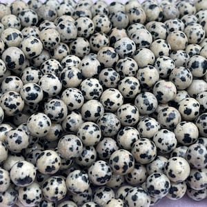 May include: A close-up of numerous Dalmatian jasper beads. Each bead is a creamy white colour, speckled with black spots. The beads are spherical and appear to be smooth, likely intended for use in jewellery or other craft projects.