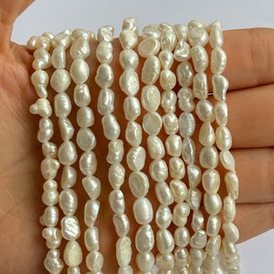 May include: A strand of white freshwater pearls, with a baroque shape, are strung together on a white thread. The pearls are irregular in shape and size, and have a smooth, glossy finish.
